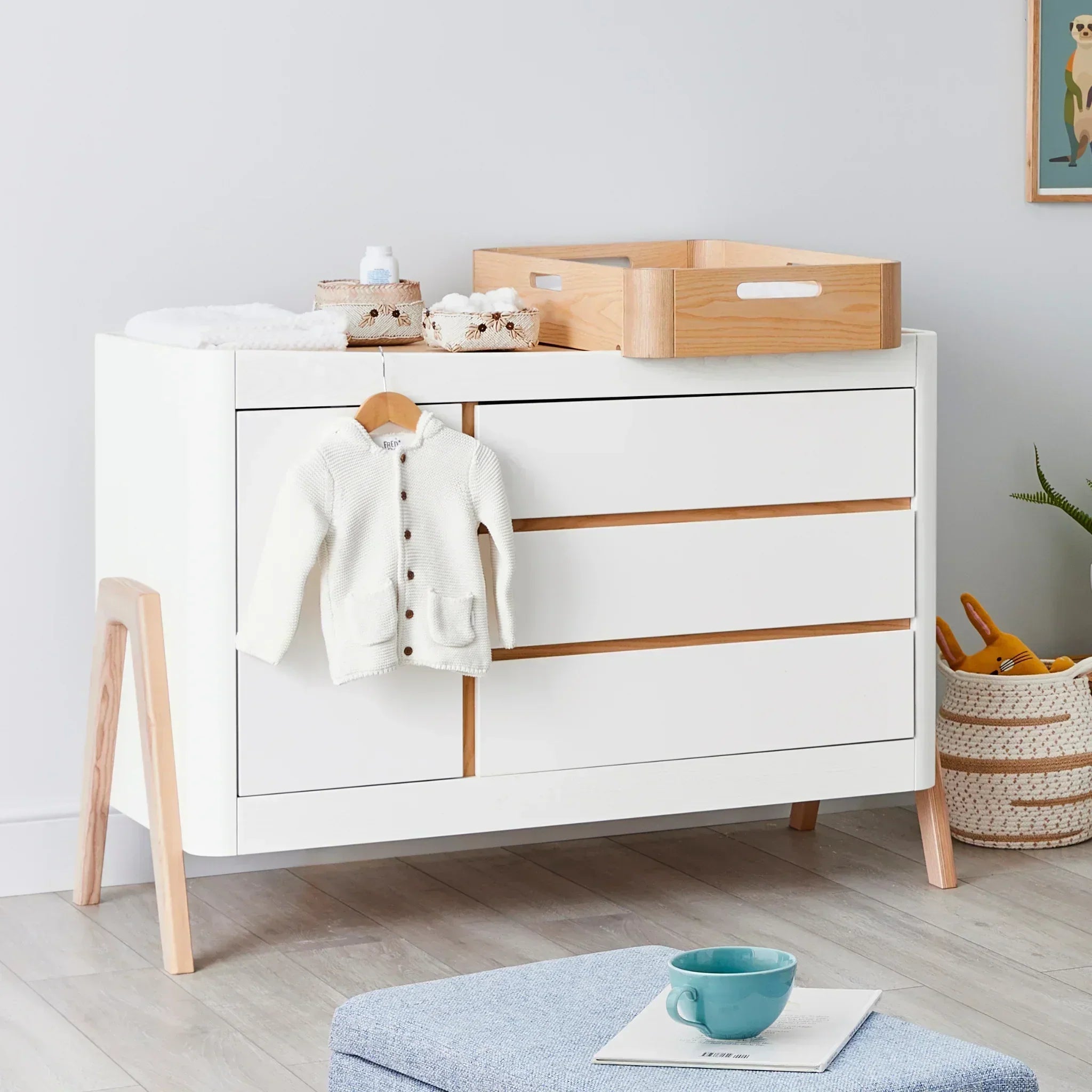 Gaia Baby Hera Dresser in white and natural wood with the Hera Changing Station placed on top, styled with baby essentials and a hanging knitted cardigan in a modern nursery.