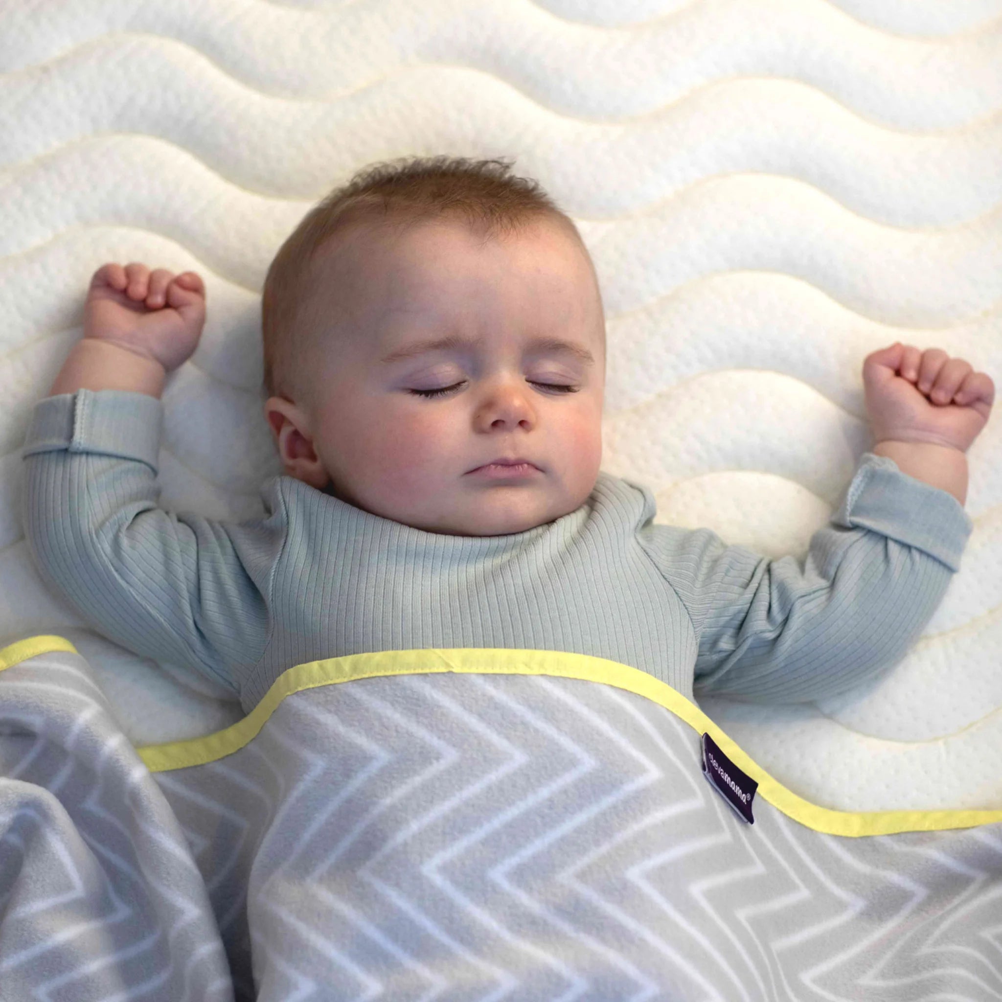 Bébé dort paisiblement sur le matelas de luxe à ressorts ensachés ClevaFoam de ClevaMama, recouvert d'une couverture douce à motifs.