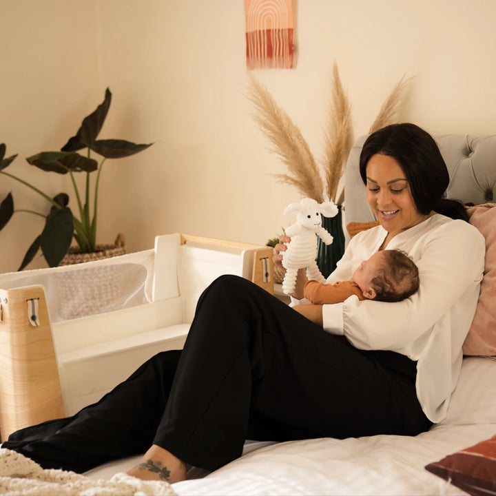 Mum playing with her baby and a teddy with the Hera bedside crib next to her bed.