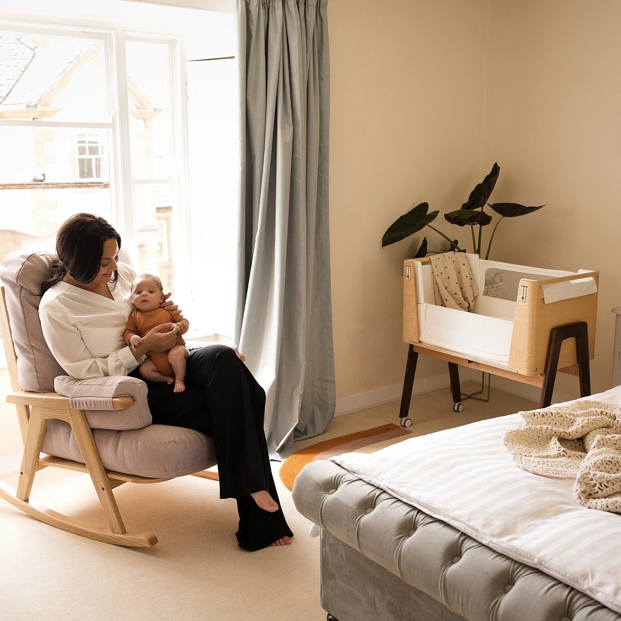 Mum sitting on Hera rocking chair with baby on her knee and the Hera Bedside Crib is in the corner of the room.