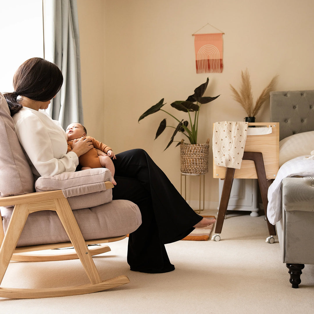 Mum holding baby on her knee on the Hera rocking chair with bedside crib next to the parents bed.
