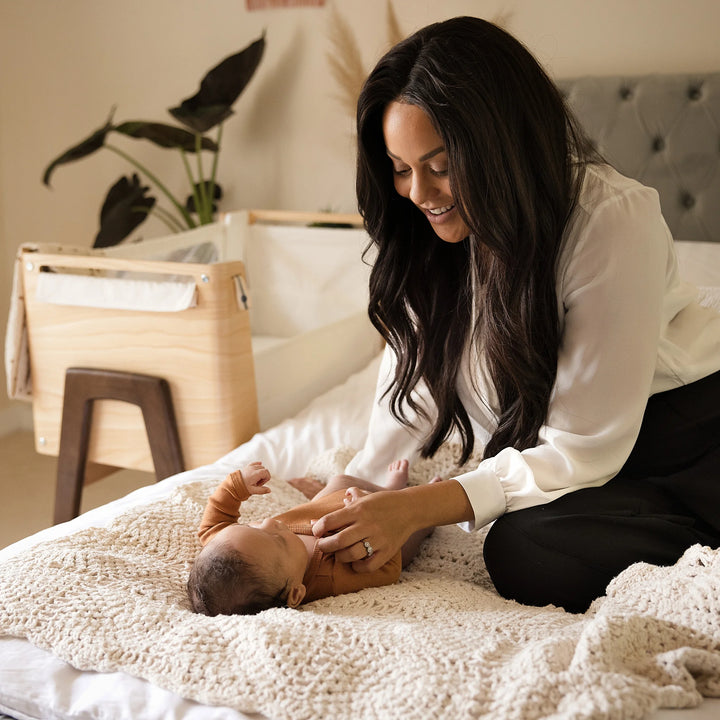 Mum playing with baby on the bed with the real wood Hera bedside crib in the background.