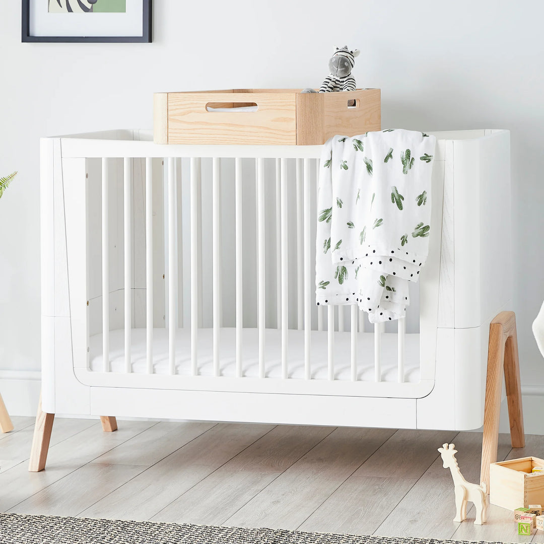 Gaia Baby Hera Changing Station in Natural Wood atop the Gaia Baby Hera Cot Bed in Scandi White, styled in a modern nursery with soft toy and blanket.