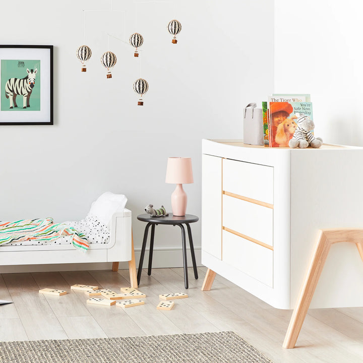Gaia Baby Hera Dresser in scandi white in a modern with soft toys and books on top. The Gaia Baby Hera Cot Bed in first bed mode partially visible to the side.