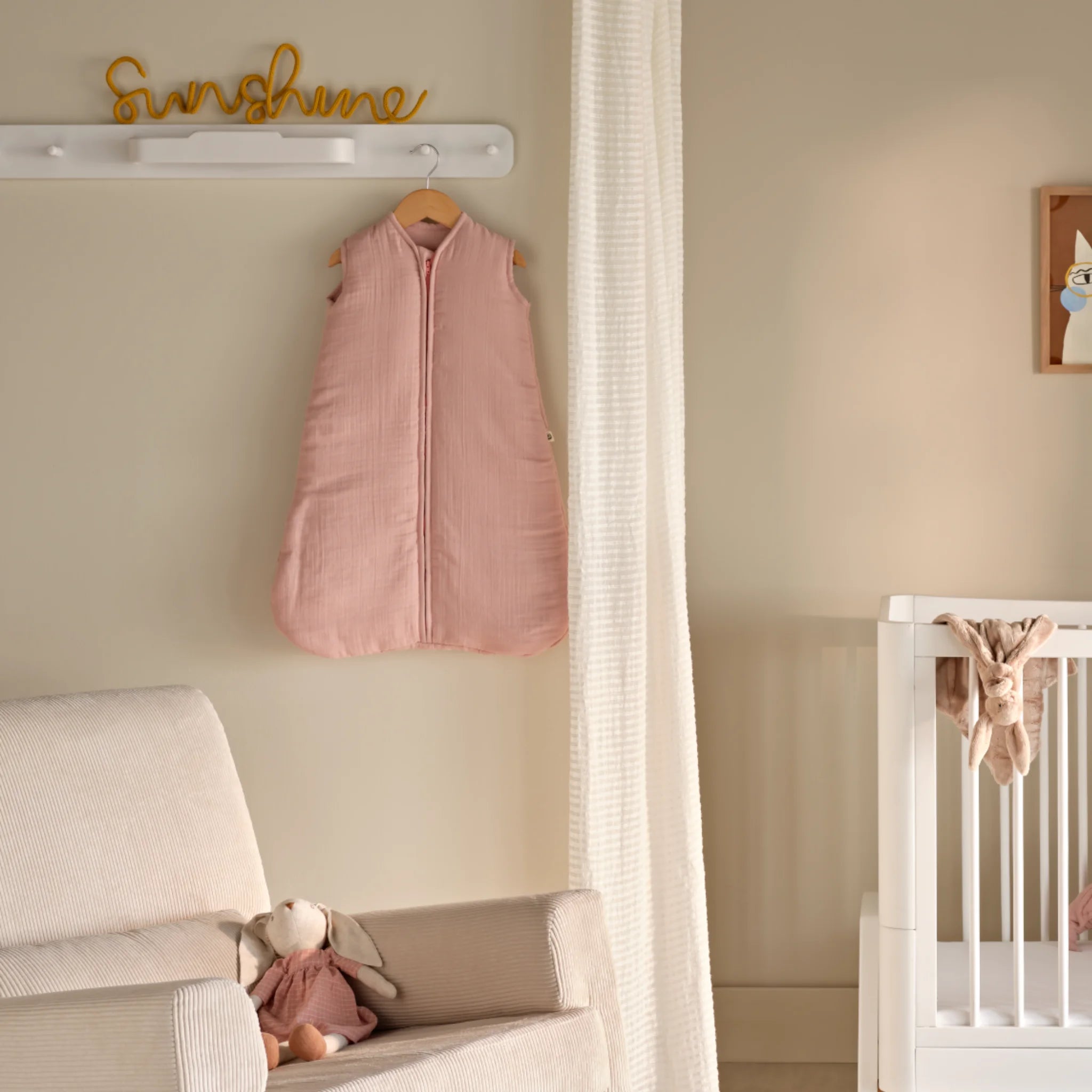Hera weiß hängende Kleiderstange an der Wand des Kinderzimmers, mit Maia ärmellosem Musselin-Schlafsack in Rosa, der an einer Wäscheklammer über einem hellbeigen/ecrufarbenen Schaukelstuhl mit einem kleinen Teddybären hängt. Rechts im Bild ist das Ende eines weißen Holzbetts von Hera Studio Edit zu sehen.
