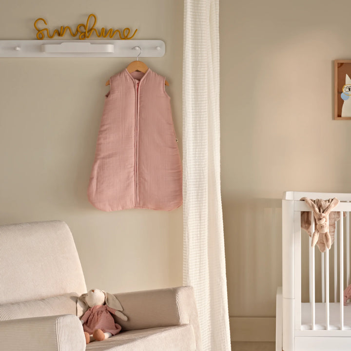 Hera white hanging peg rail on nursery wall with Maia muslin sleeveless sleeping bag in pink hanging on a peg above a pale beige / ecru rocking chair with a small bunny teddy. There is the end of a white Hera Studio Edit wooden cot on the right of the image.