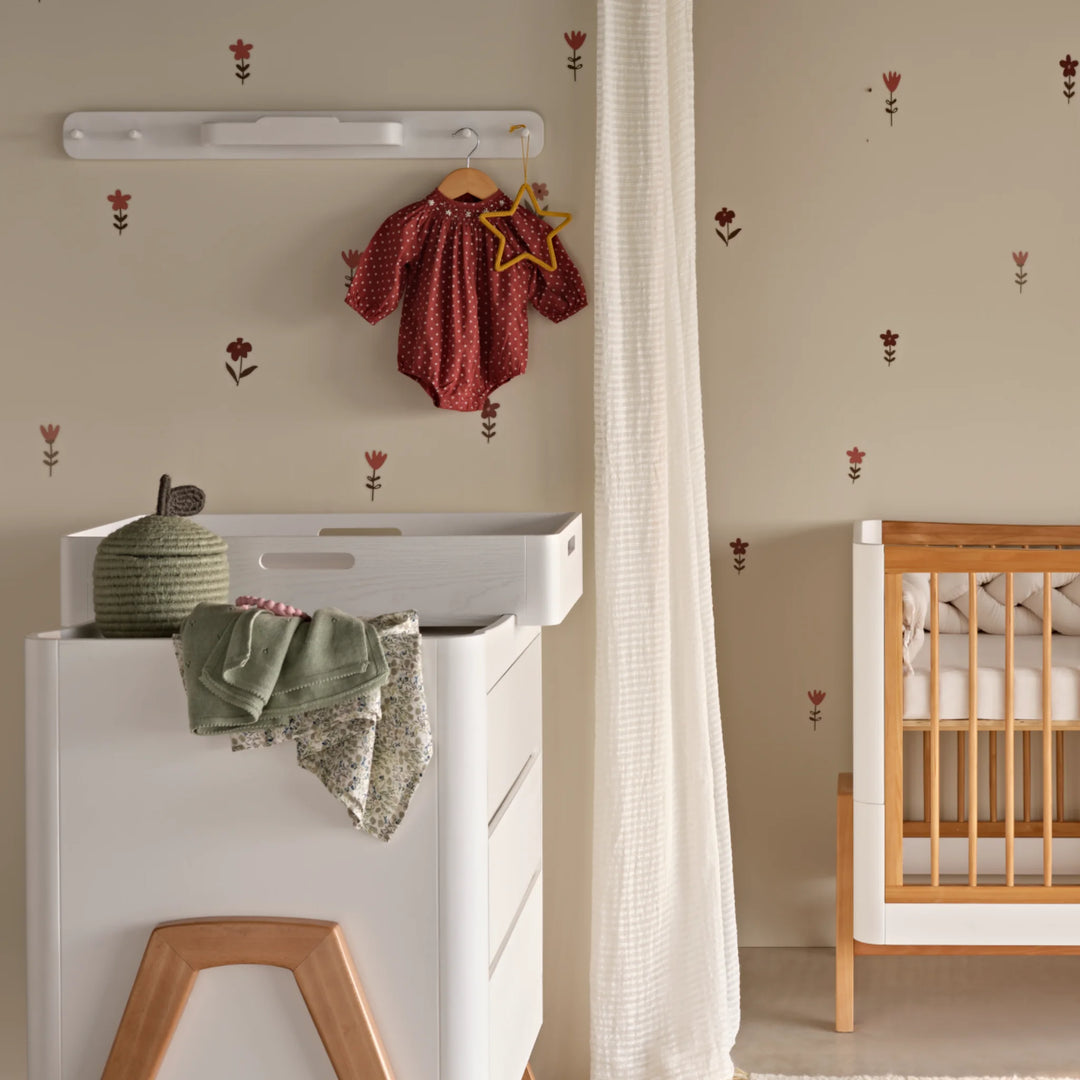 Nursery scene featuring the white Hera peg board hanging rail on the wall above a matching white dresser with changing top. There are baby clothes on the dresser and a vest hanging on one of the pegs above.