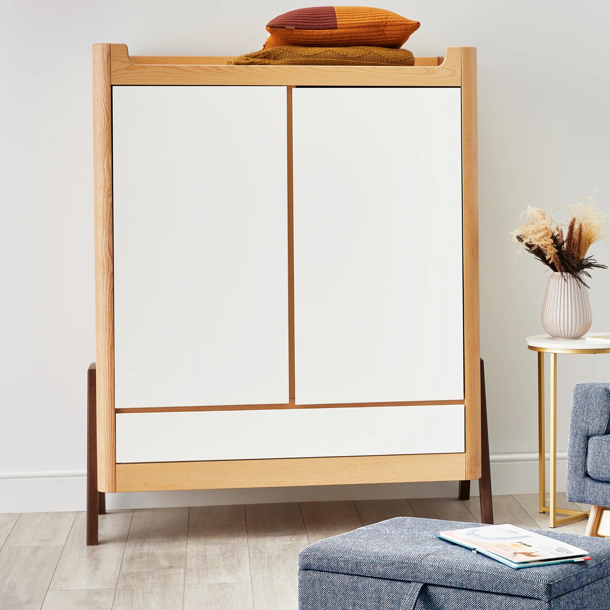 Armoire Gaia Baby Hera en bois naturel blanc dans une chambre de bébé moderne et minimaliste avec coussins et couvertures rangés sur le dessus.
