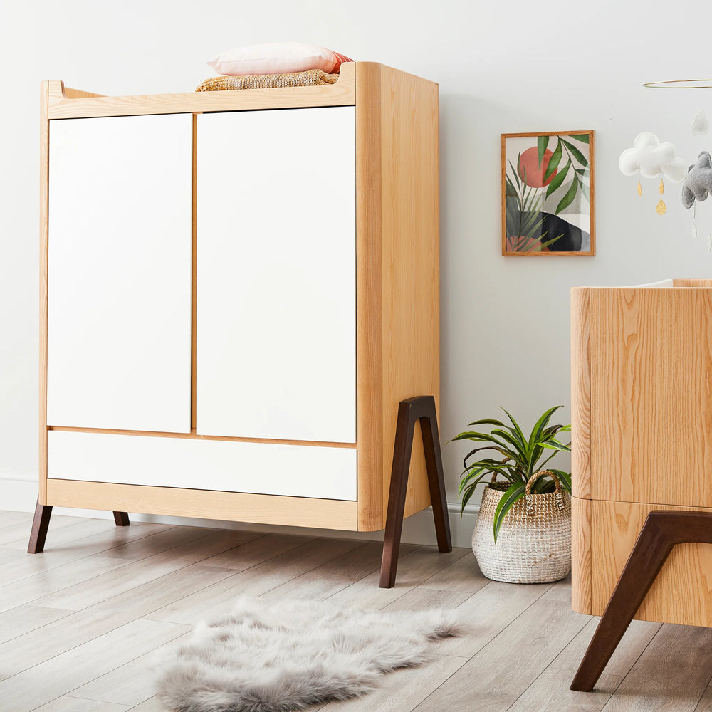 Angled view of Gaia Baby Hera Wardrobe in white natural wood with walnut a-frame legs in a modern nursery with Gaia Baby Hera Cot Bed partially visible.
