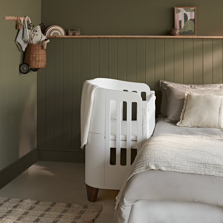 Gaia Baby Serena Bedside Crib Add On with walnut legs side shot of it beside a parents bed