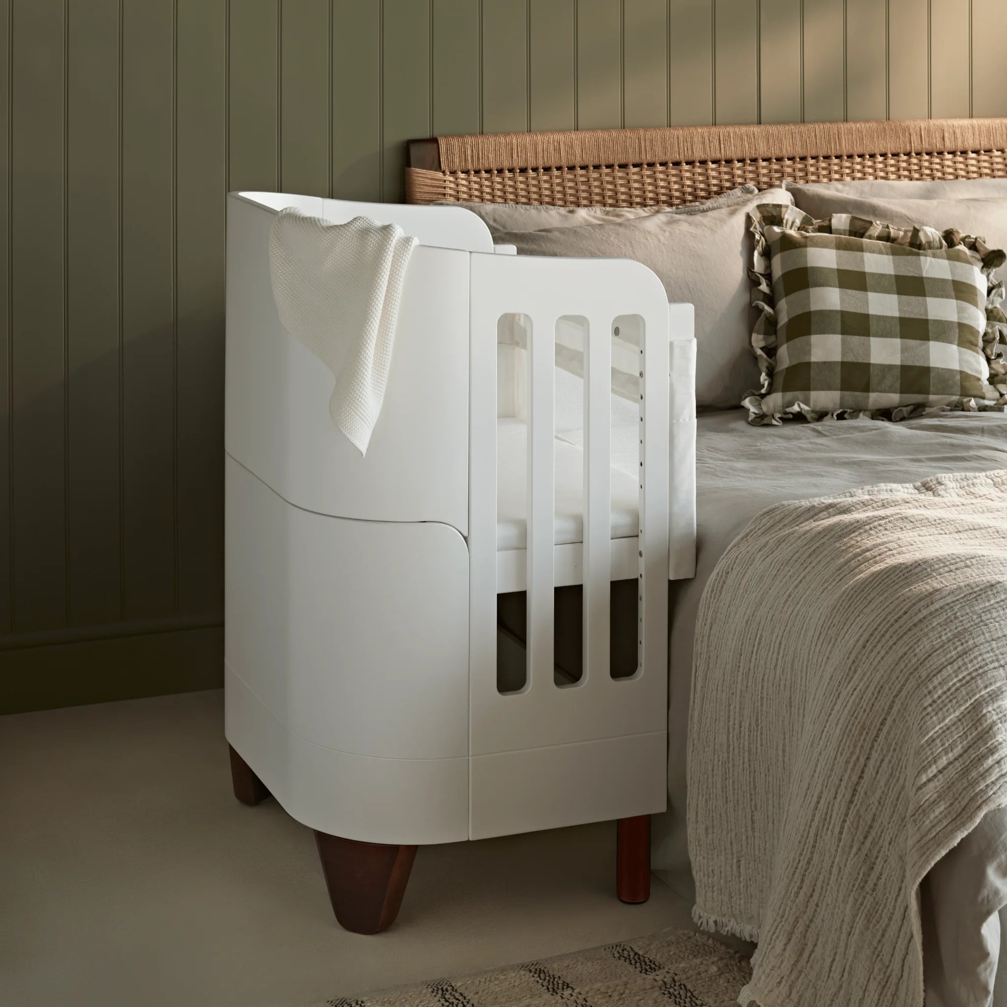 Gaia Baby Serena Beistellbett mit Walnussholzbeinen neben einem modernen Bett, stilvolle Beistellwiege für Neugeborene in einer warmen, neutralen Schlafzimmerumgebung mit strukturierter Bettwäsche und sanfter Umgebungsbeleuchtung.