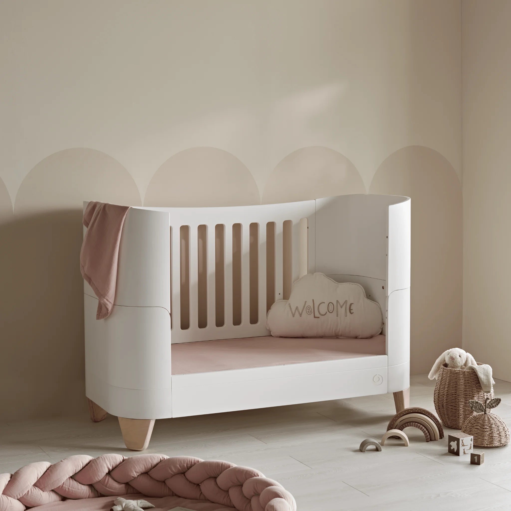 Gaia Baby Serena Kinderbett im Kleinkinderbettmodus mit Beinen aus Naturholz, umwandelbares weißes Kleinkinderbett für modernes Kinderzimmer und Babyzimmer im skandinavischen Stil mit zartrosa Dekor.