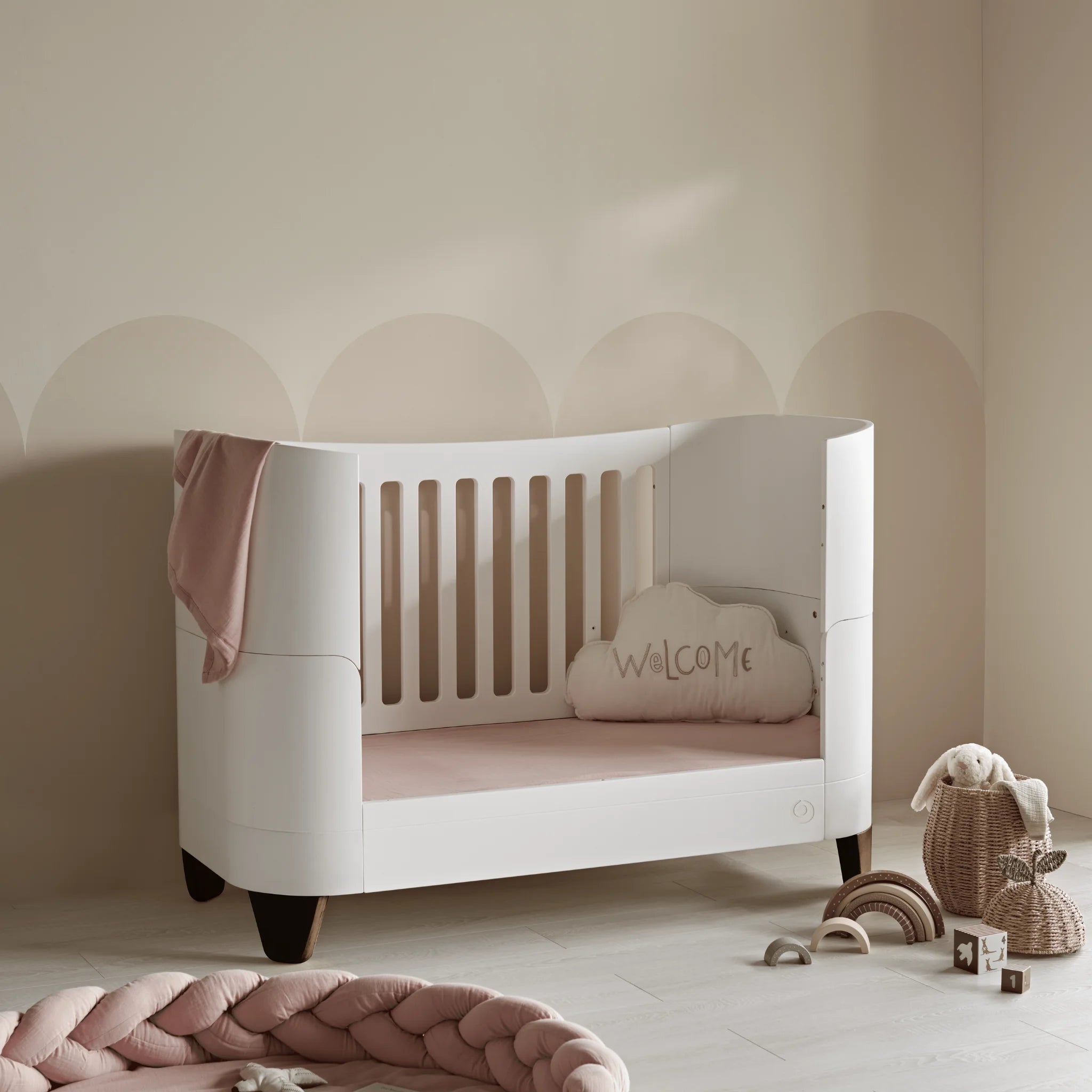 Gaia Baby Serena Kleinkinderbett mit Beinen aus Walnussholz in einem weichen, neutralen Kinderzimmer, modernes weißes Kleinkinderbett mit rosafarbener Bettwäsche, Regenbogenspielzeug, geflochtenen Körben und gemütlicher, moderner Kinderzimmerdekoration.