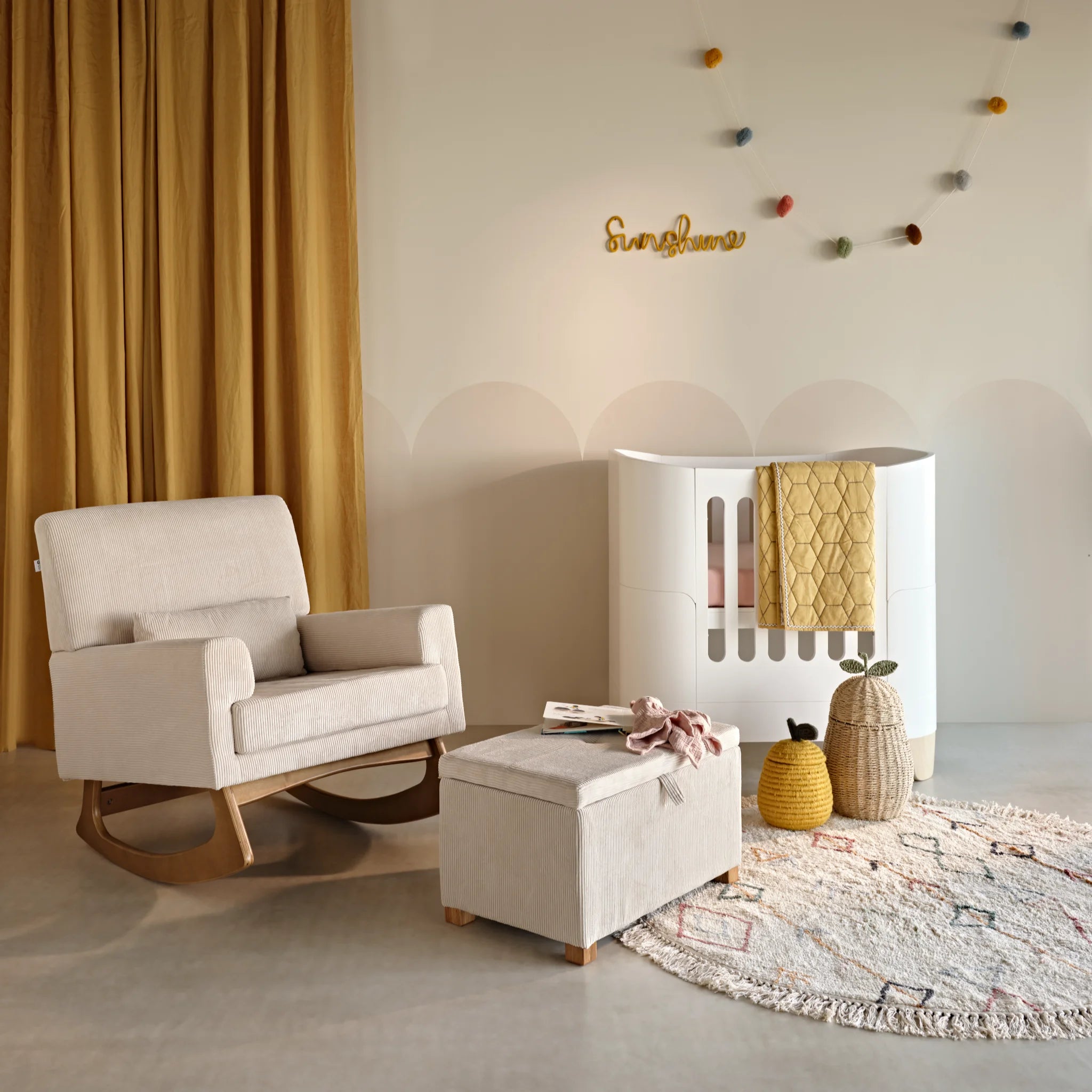 Warm nursery scene featuring a white Serena mini cot beside an ecru rocking chair and matching ottoman, with soft textiles, colourful garland decor, and a patterned rug.
