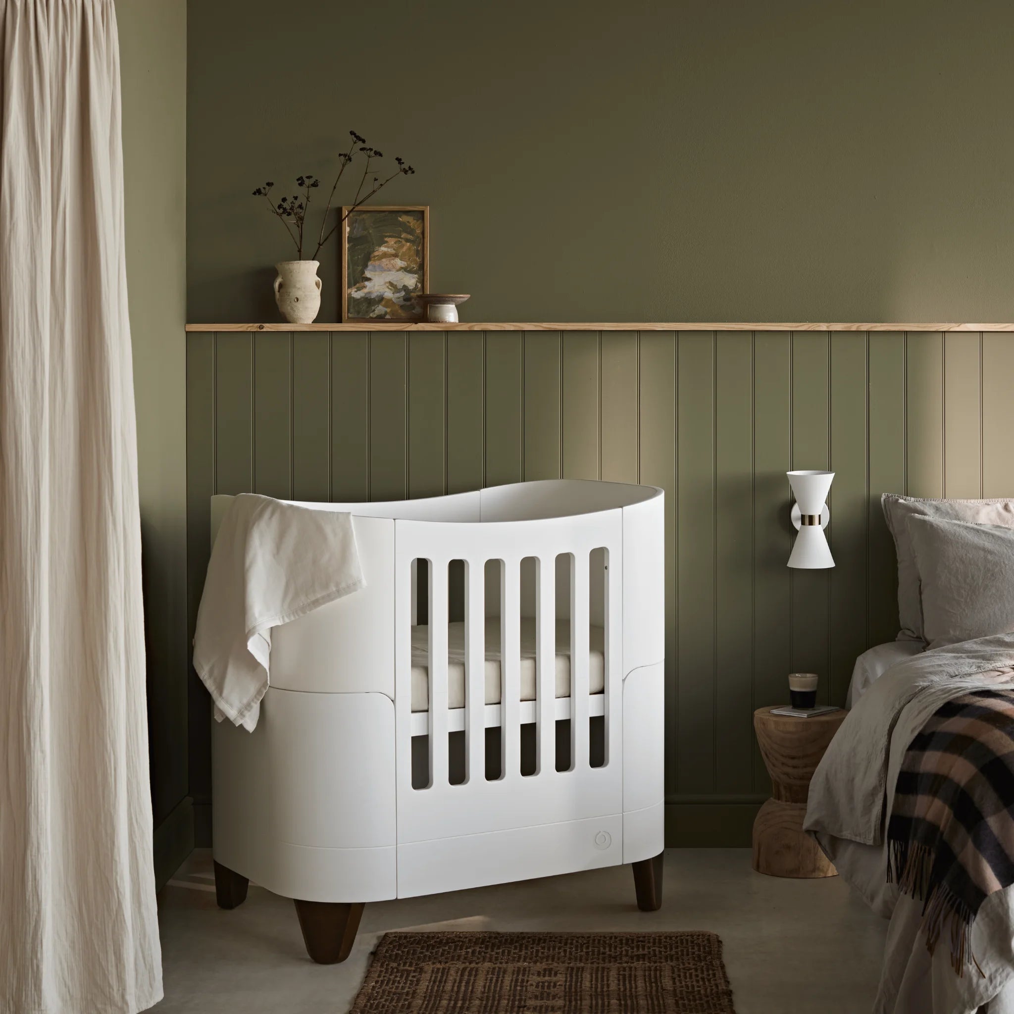 Gaia Baby Serena Mini-Kinderbett mit Walnussbeinen, weißes Babybett im stilvollen Kinderzimmer mit grün getäfelten Wänden und neutralem Dekor.