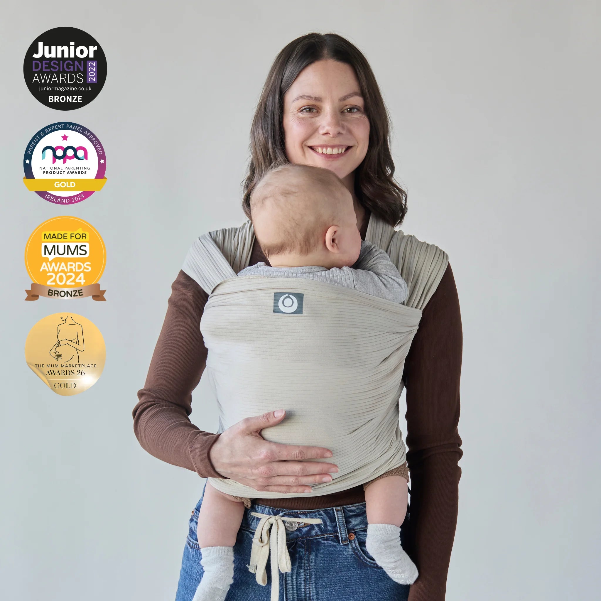Mother wearing an ecru corduroy Gaia Baby stretchy wrap holding a baby, with award badges displayed on a light background.