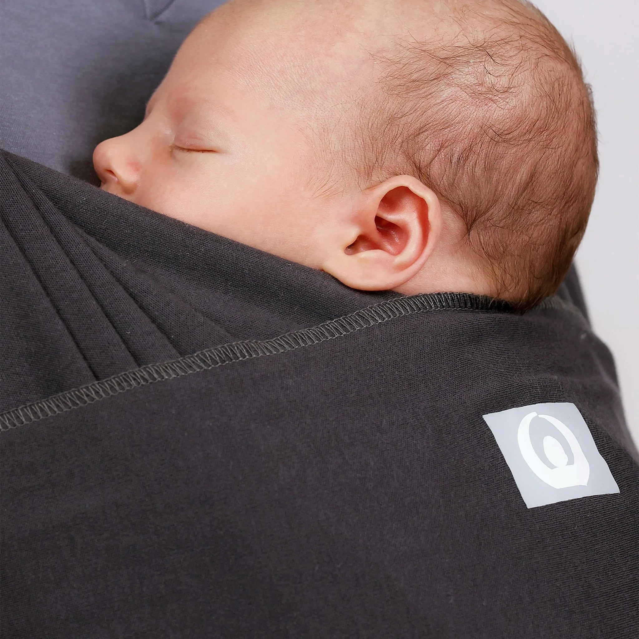 Close-up of a baby sleeping in a graphite wrap carrier with logo visible.
