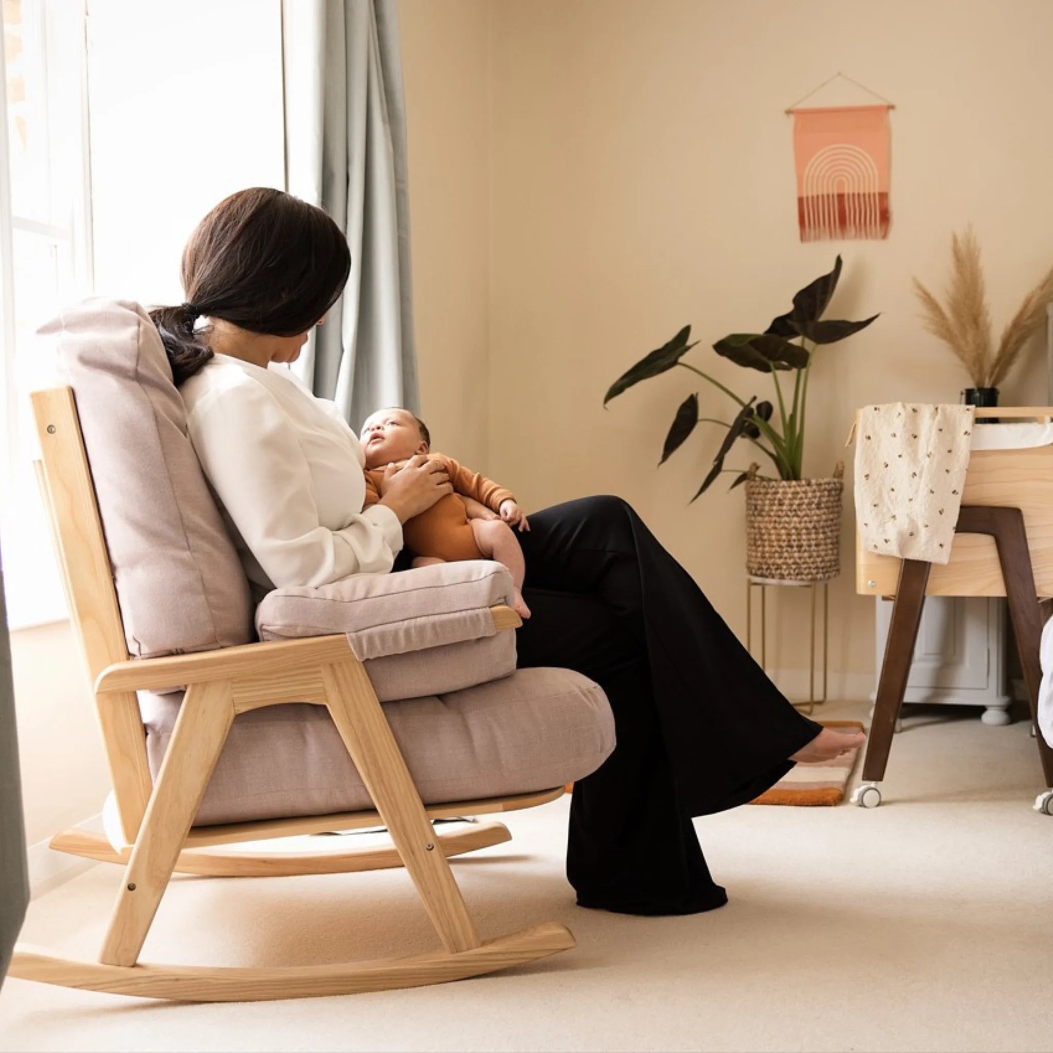 Gaia Baby Hera Rocking Chair in natural oak with blush pink cushions, modern wooden nursery glider chair for breastfeeding and newborn bonding, mother holding baby in Scandinavian style nursery room.