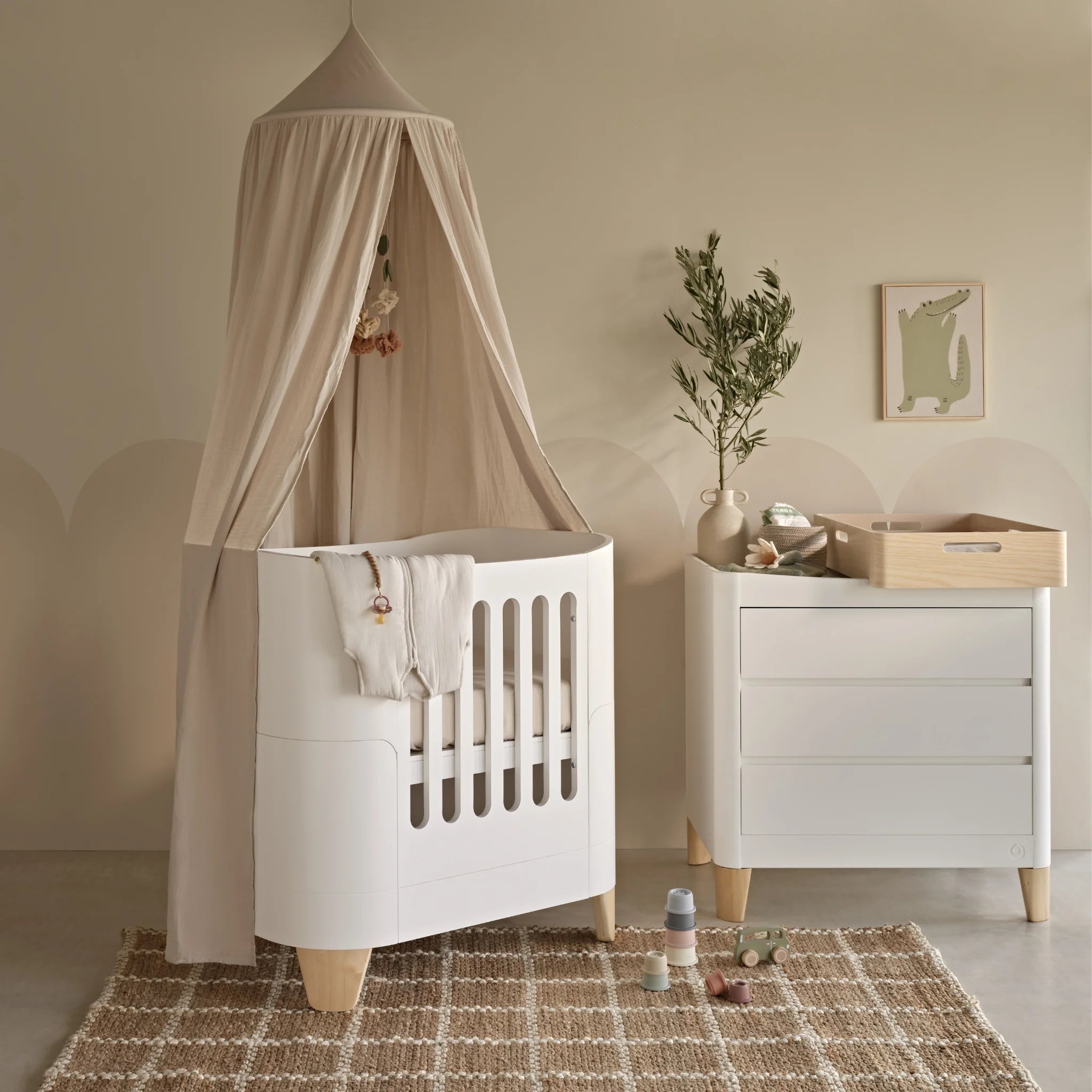 White Gaia Baby Serena mini cot with slatted sides and natural wood legs, styled with a soft fabric canopy, paired with a matching Serena dresser and Hera changing station, arranged in a warm nursery setting with neutral décor, a rug, and baby accessories.
