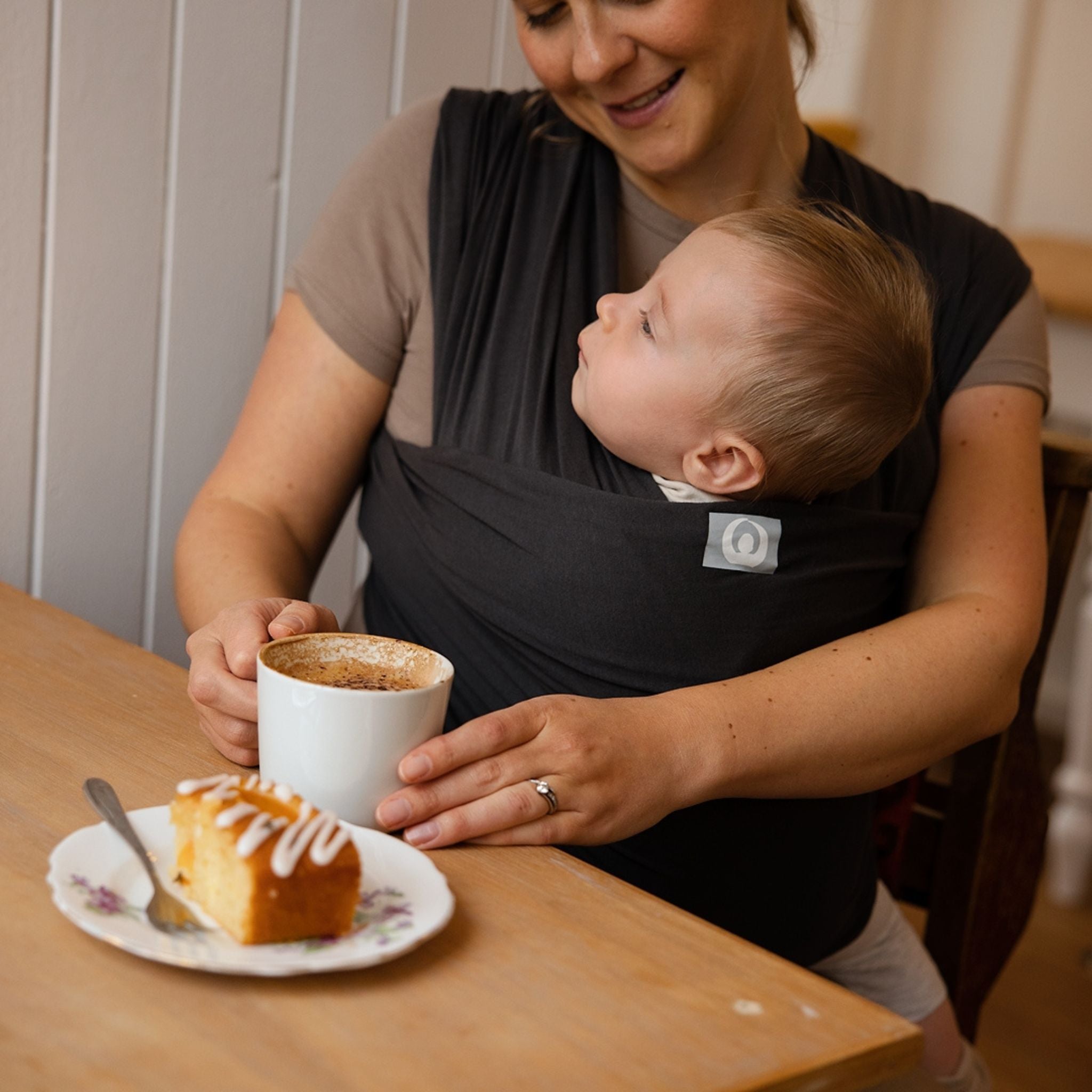 Mother enjoying coffee with baby in Gaia Baby Graphite organic cotton stretchy baby wrap carrier