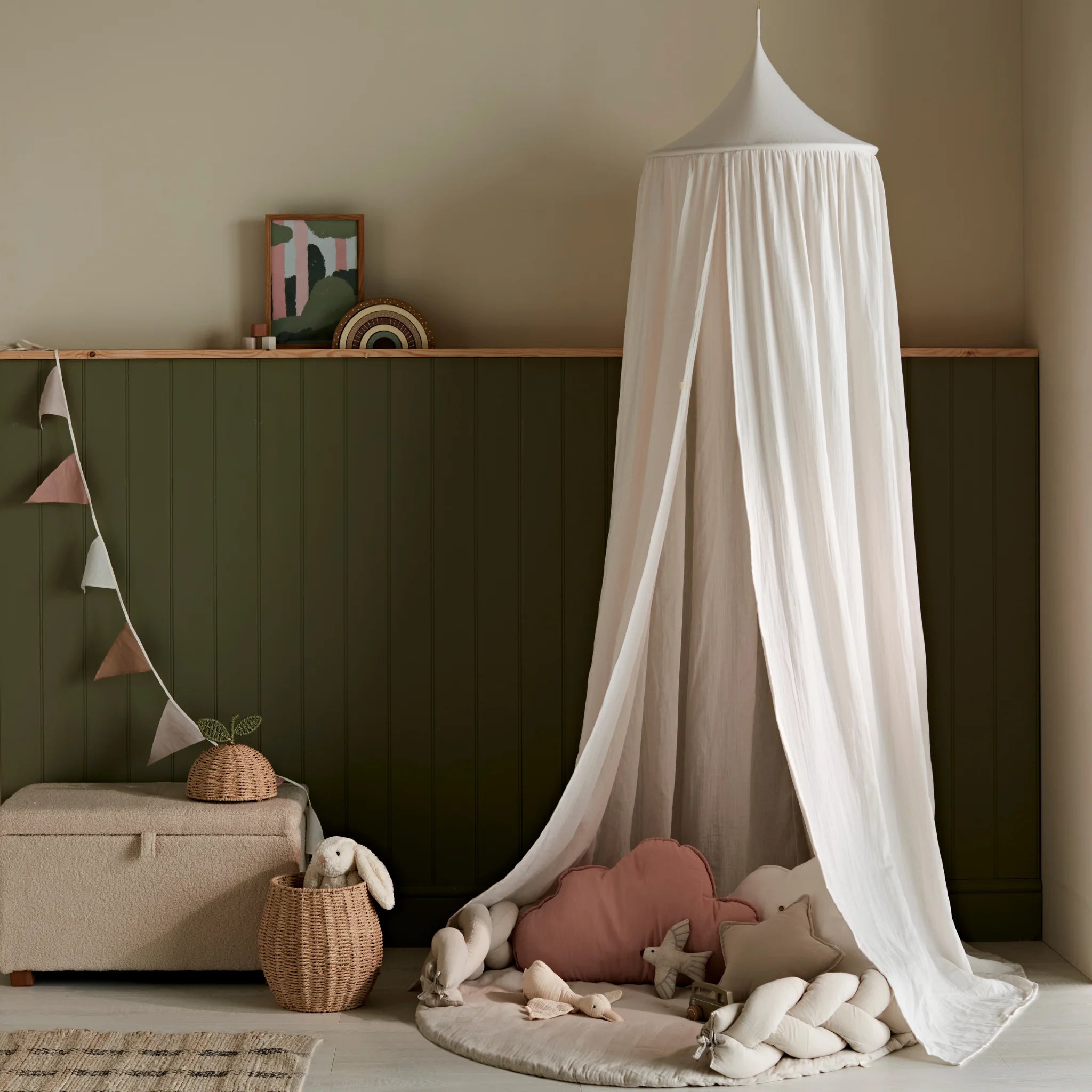 Gemütliche Kinderzimmer-Spielecke mit einem weichen weißen Baldachin, der über einer runden Spielmatte mit geflochtenen Kissenkissen, Stofftieren, geflochtenen Körben und gedämpften rosa Kissen vor olivgrün getäfelten Wänden drapiert ist.