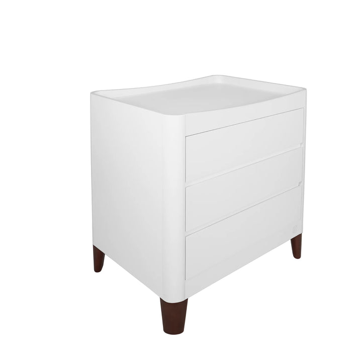 Angled view of the white Serena dresser with walnut legs, featuring three drawers and a curved changing top, displayed on a white background.
