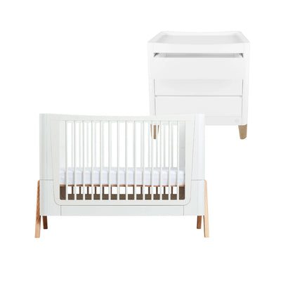 White Gaia Baby Hera cot bed with white slatted sides and light natural wood legs, paired with a matching white Serena changing dresser, displayed on a white background.