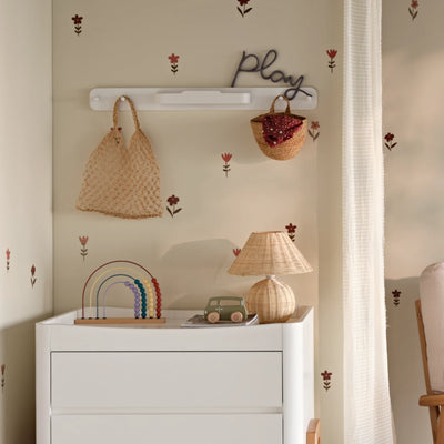 White Hera hanging peg rail on the wall of a nursery with a mesh bag and woven basket above a matching white Hera nursery dresser with toys on top.