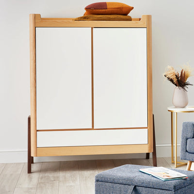 Armoire Gaia Baby Hera en bois naturel blanc dans une chambre de bébé moderne et minimaliste avec coussins et couvertures rangés sur le dessus.