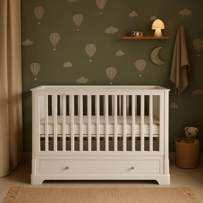 White cot in a room with green walls featuring hot air balloons and clouds, a teddy bear, and a lamp.