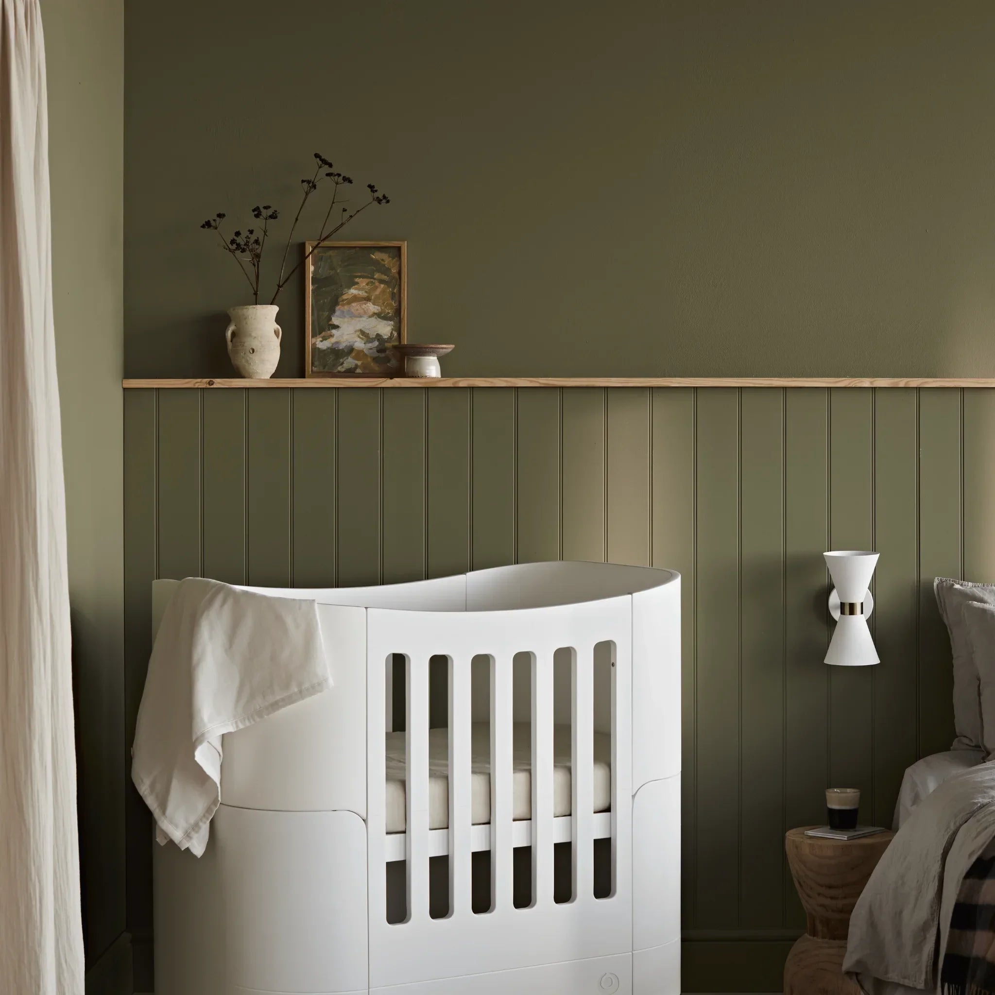 White muslin baby blanket draped over a modern white cot in a stylish nursery with dark olive green panelled walls and soft ambient lighting.