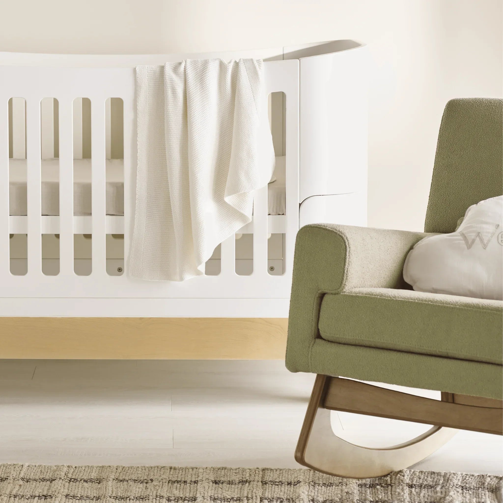 Soft white muslin baby blanket draped over the side of a white cot in a calm neutral nursery setting, with part of a green upholstered rocking chair visible in the foreground.