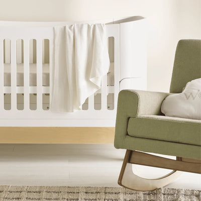 Weiche, weiße Musselin-Babydecke drapiert über der Seite eines weißen Kinderbetts in einer ruhigen, neutralen Kinderzimmerumgebung, mit einem Teil eines grün gepolsterten Schaukelstuhls im Vordergrund.