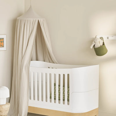 Neutrale Kinderzimmerecke mit einem weißen Kinderbett unter einem weichen weißen Musselin-Baldachin, gestaltet mit natürlichen Holzakzenten, gedämpfter grüner Bettwäsche und minimalistischer Wanddekoration für eine ruhige und gemütliche Atmosphäre im Babyzimmer.