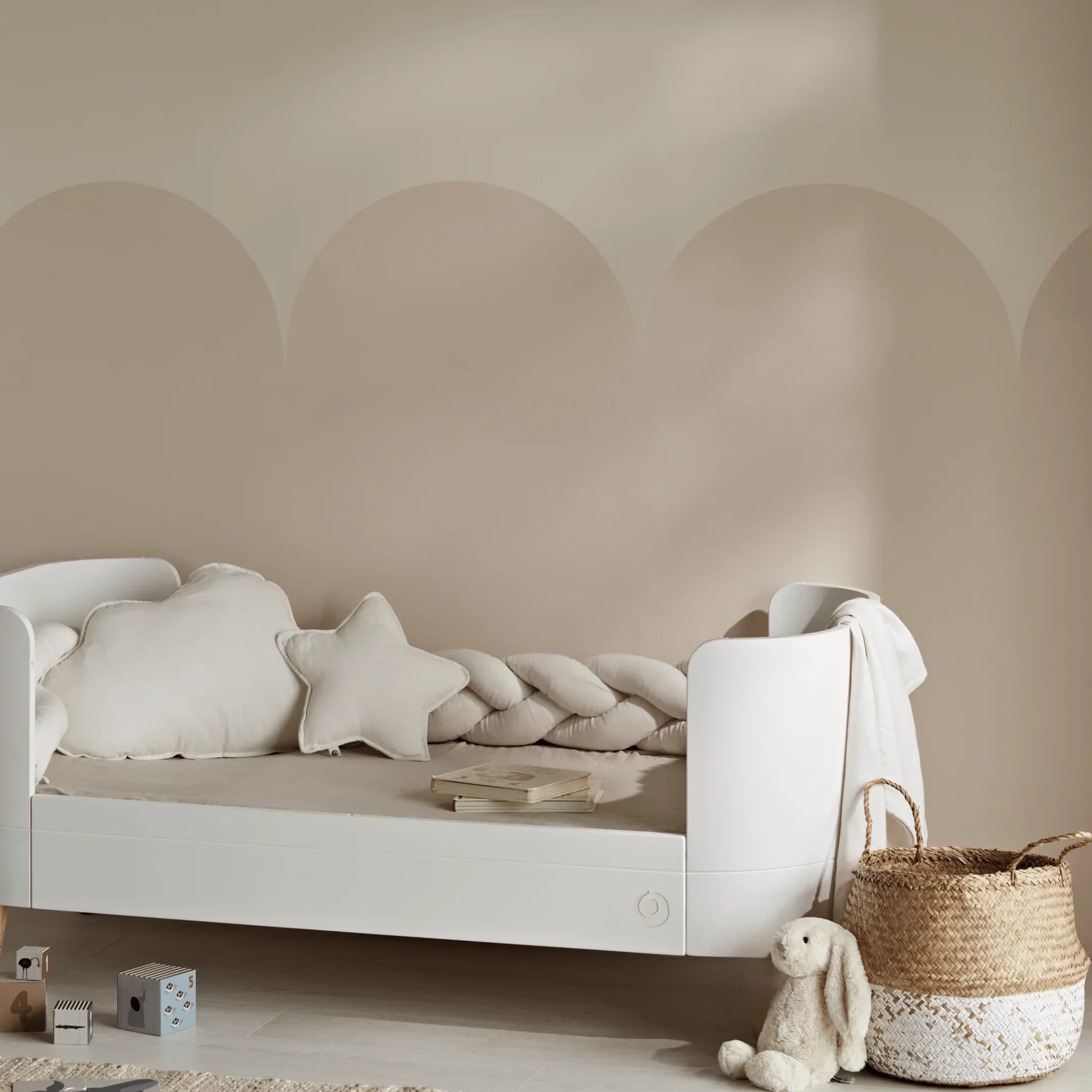 Minimalist toddler bed styled with soft white decorative cushions, including cloud and star-shaped pillows, a braided bumper cushion, woven baskets, and plush toys in a warm neutral nursery setting.