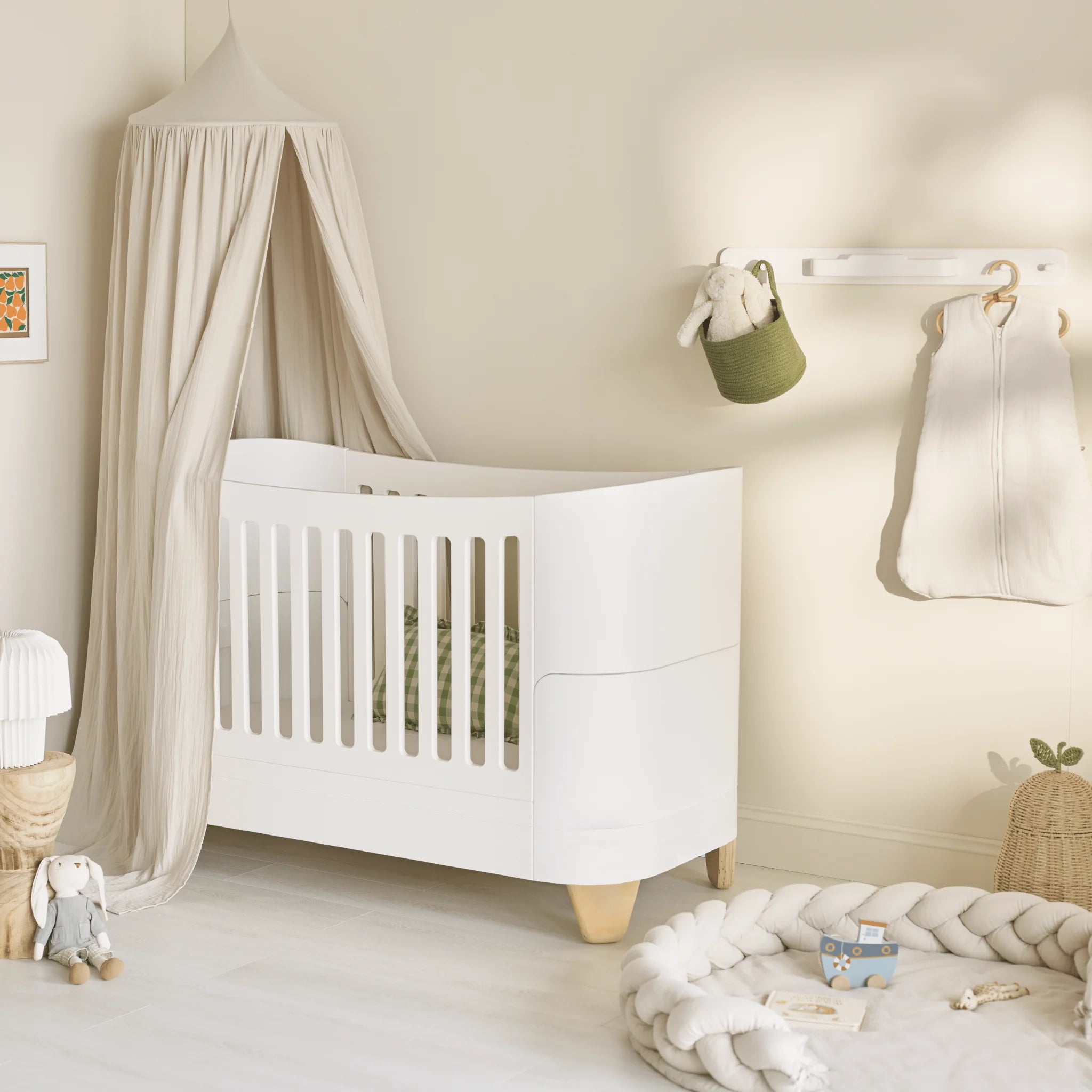 Minimalist nursery featuring a white Serena cot bed with natural wood legs, styled beneath a soft beige canopy beside a braided play mat and neutral-toned nursery accessories.