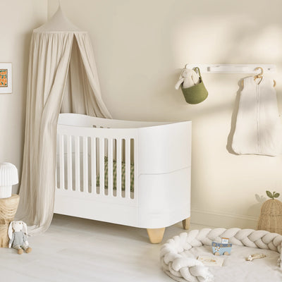 Minimalist nursery featuring a white Serena cot bed with natural wood legs, styled beneath a soft beige canopy beside a braided play mat and neutral-toned nursery accessories.