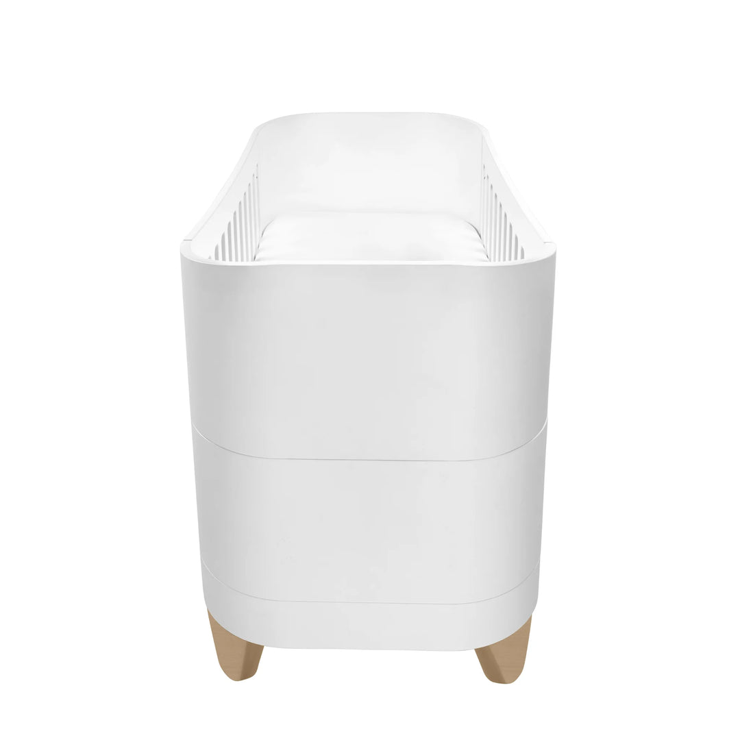 Gaia Baby Serena cot bed with natural wood legs shown from the side, modern white convertible baby crib with curved edges and minimalist Scandinavian nursery design on a clean white background.