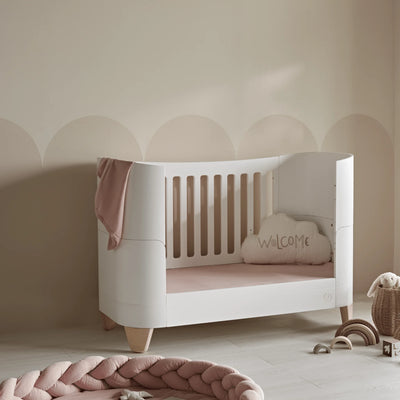 Modern white toddler cot bed with natural wood legs, blush pink fitted sheets and decorative nursery cushion in a soft neutral baby room.