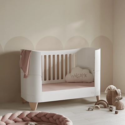 Gaia Baby Serena Kinderbett im Kleinkinderbettmodus mit Beinen aus Naturholz, umwandelbares weißes Kleinkinderbett für modernes Kinderzimmer und Babyzimmer im skandinavischen Stil mit zartrosa Dekor.