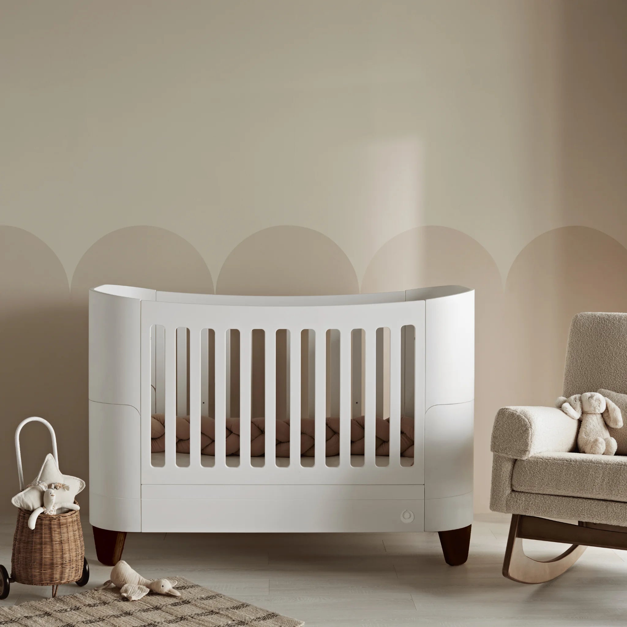 Serena cot bed with walnut legs styled in a warm neutral nursery setting beside a boucle rocking chair, woven storage basket, and soft toys, creating a calm and contemporary space for baby.