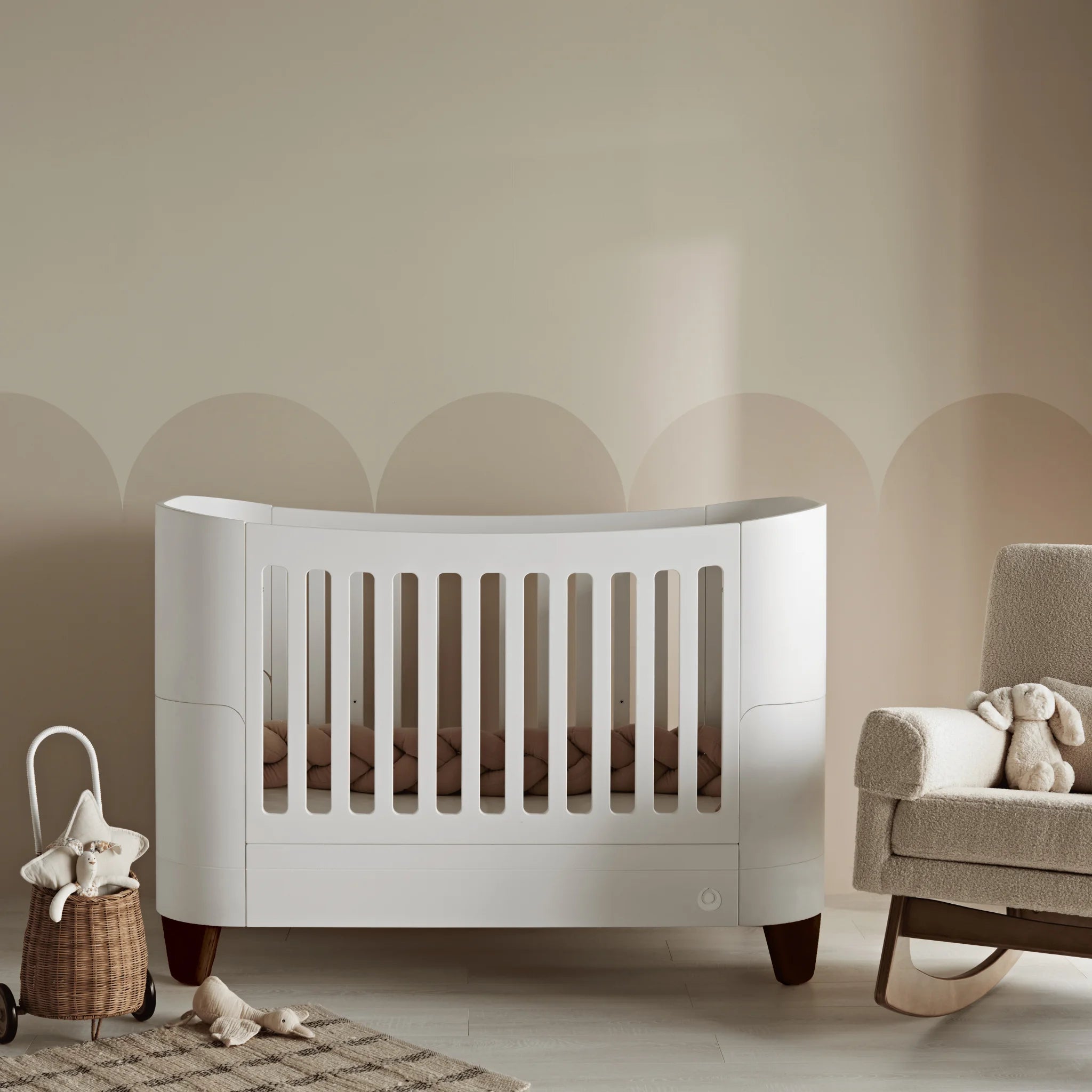 Gaia Baby Serena Kinderbett mit Beinen aus Walnussholz in einem ruhigen, neutralen Kinderzimmer, modernes weißes Babybett mit sanften Beigetönen, geflochtenem Korbdekor und passender Schaukelstuhl in einem warmen, modernen Babyzimmer.