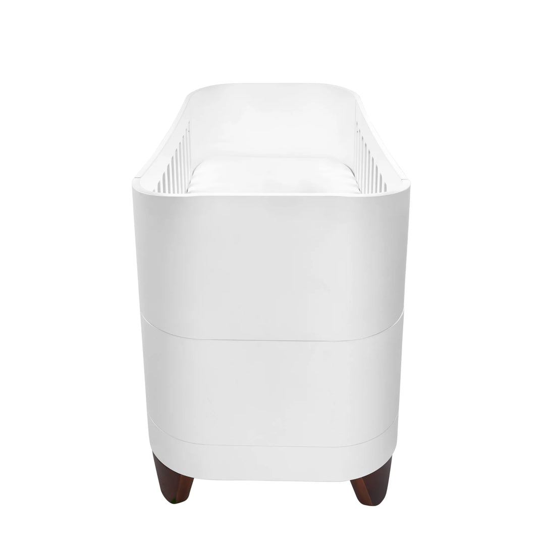 Side view of the white Serena cot bed with walnut legs, showing the curved end panels and slatted interior, displayed on a white background.