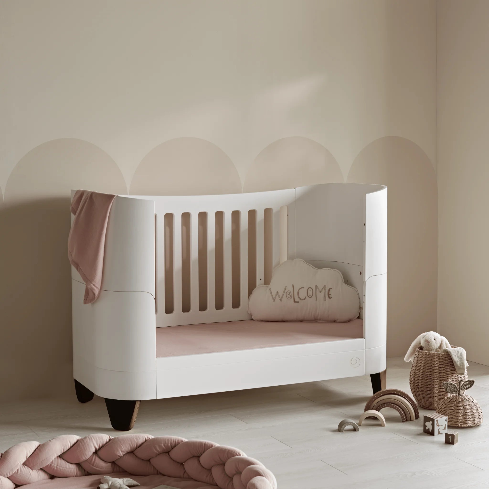 Gaia Baby Serena Cot Bed in toddler bed mode with walnut legs, white wooden toddler bed with soft pink bedding in neutral nursery.