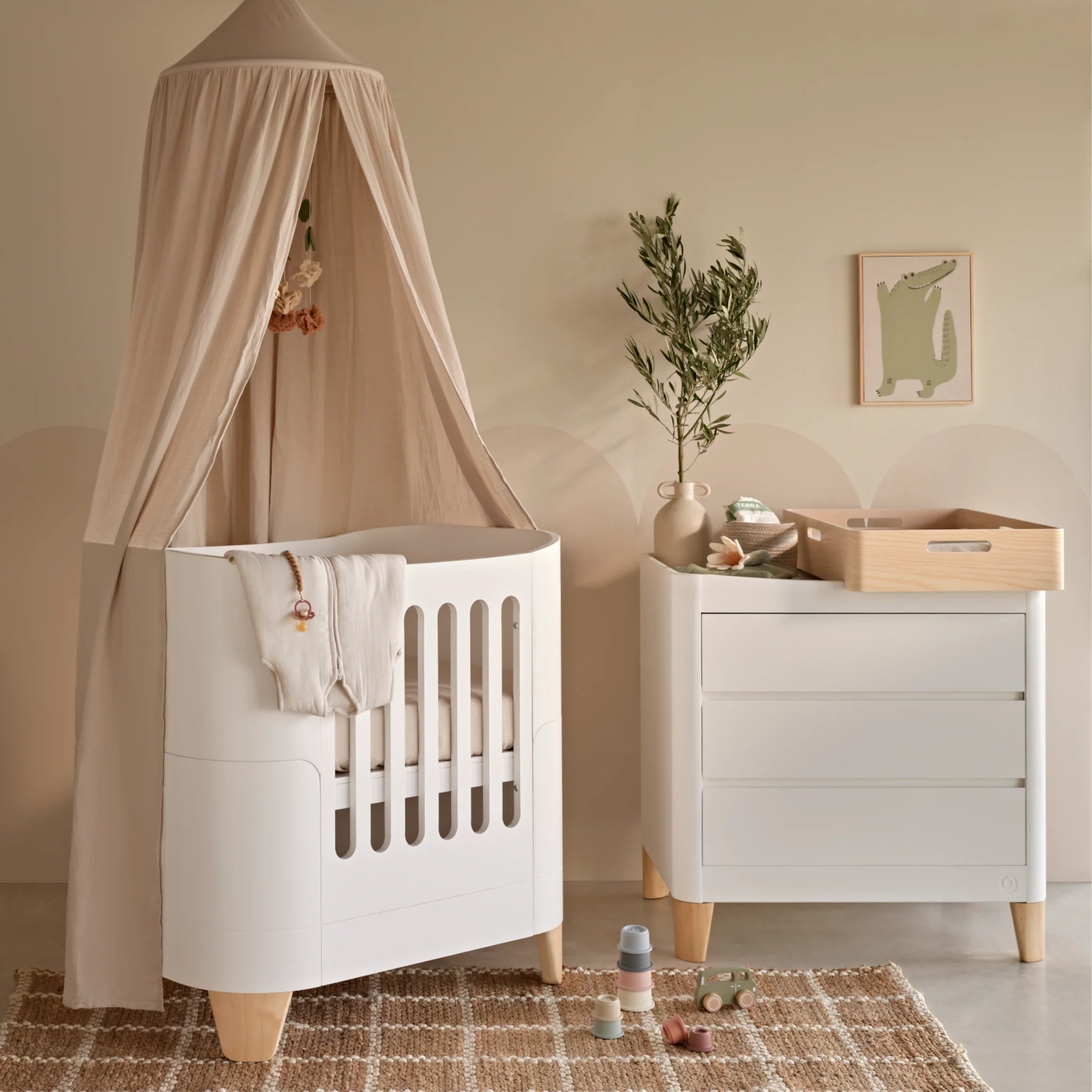 Serena Mini Cot and Dresser with natural legs and maia muslin canopy. The Hera changing station in natural is on top of the dresser.