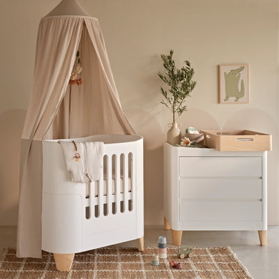 Serena Mini Cot and Dresser with natural legs and maia muslin canopy. The Hera changing station in natural is on top of the dresser.