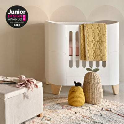 Gaia Baby Serena Mini Cot in Natural and Ecru with wooden legs, styled in a nursery with a mustard quilt draped over the side, woven storage baskets, and a textured rug. Junior Design Awards 2024 Gold winner badge displayed.