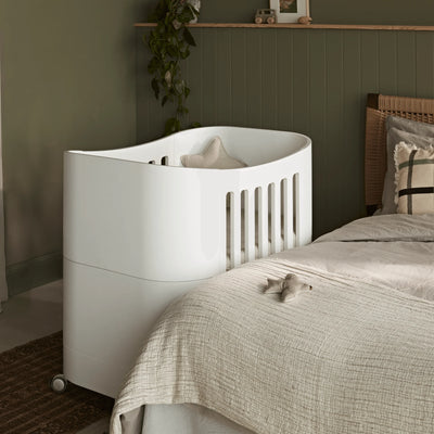 Weißes Beistellbett mit Rollen neben einem Bett in einem gemütlichen Schlafzimmer mit dunkelgrün getäfelten Wänden, neutraler Bettwäsche und sanfter Umgebungsbeleuchtung.