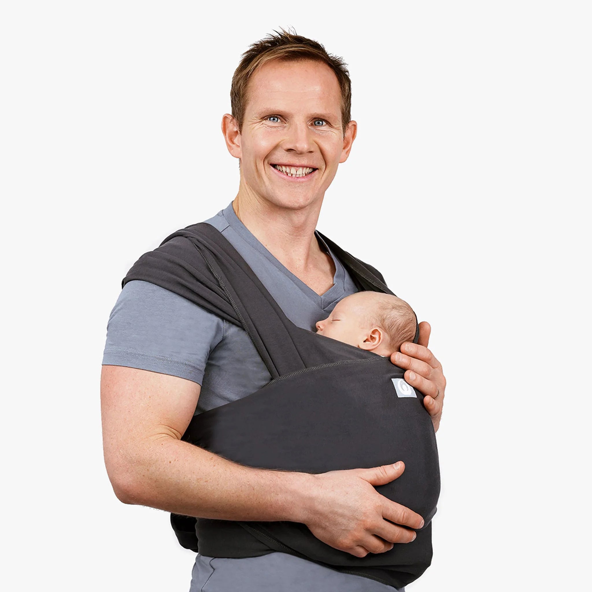 Man holding a baby in a graphite wrap carrier against a plain background.
