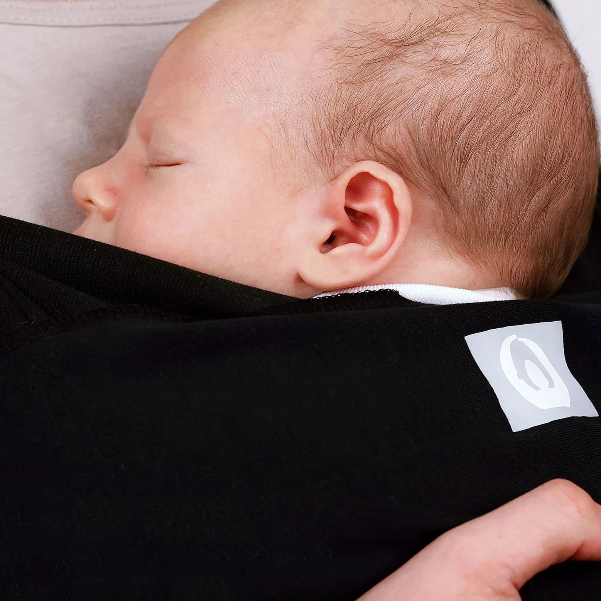 Close-up of a sleeping baby in a black Gaia Baby stretchy wrap with logo label visible.